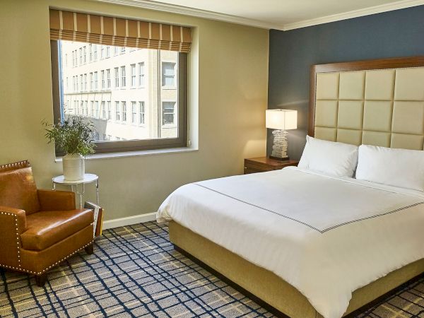 A modern hotel bedroom with a large tufted bed, bedside lamp, lounge chair, and a window with blinds.