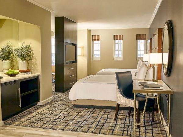 A stylish hotel room with a bed, desk, large windows with shades, and a small bar area with plants.