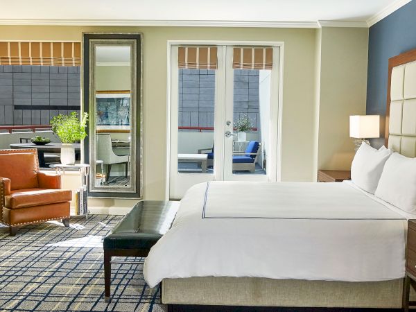 Modern hotel bedroom with a large bed, lamp, brown leather chair, and a tall mirror reflecting the lounge through doors.