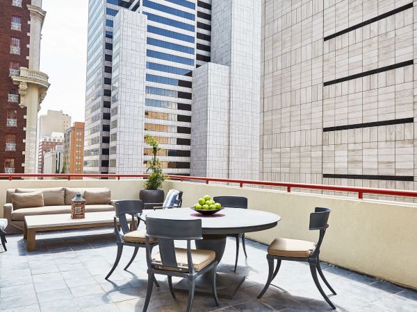 A rooftop terrace with outdoor seating and a table of fruit, facing tall city skyscrapers and brick-and-stone buildings.