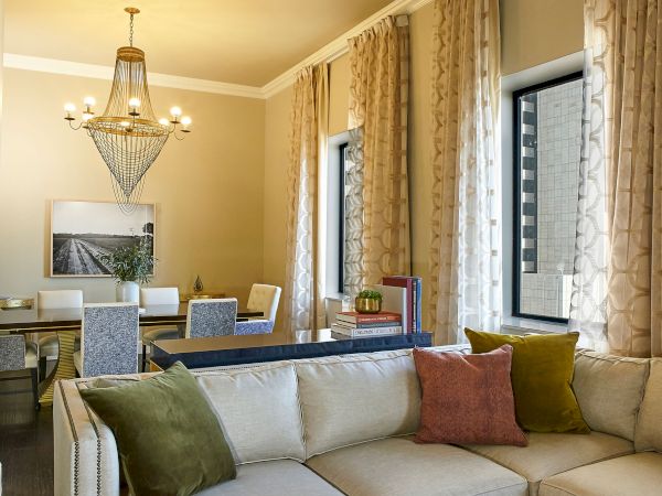 A bright living/dining room with a gold chandelier, upholstered seating, framed wall art, floor-to-ceiling curtains, and window views.