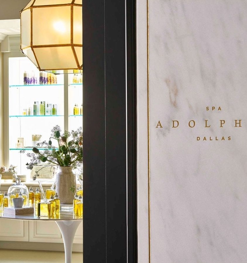 An Adolphus Dallas spa entrance with a marble wall, gold trim, and shelves of skincare oils and bottles.