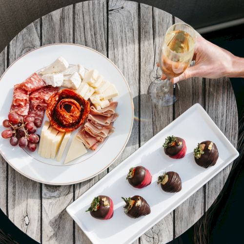 A charcuterie and cheese platter with salami, grapes, and cheeses, plus chocolate bites, while someone toasts with a glass of wine.